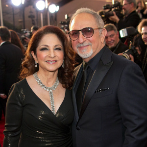 Gloria Estefan and Emilio Estefan Jr., both Caucasian, attend an awards show. Gloria in a shiny black dress with a necklace. Emilio in a tailored suit. Crowded event setting, paparazzi flashes.