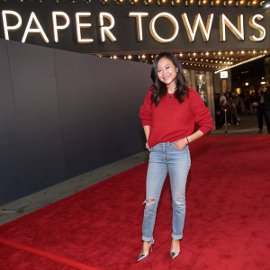 Natalie Tran at the 'Paper Towns' premiere. Wearing a casual red sweater, jeans, and silver heels. Behind her, the movie's title lights up the red carpet.