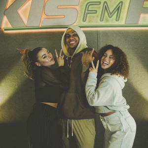 Charli XCX, Jordan Banjo, and Perri Kiely, in a vibrant studio photo. Charli in a black top, Jordan in a unique hoodie, Perri in a zipped hoodie. They pose before a bright KISS FM logo backdrop.
