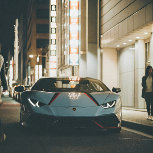 Lamborghini Huracan EVO, blue with red and black accents, parked on a bustling street at night. Neon signs, crowded cityscape