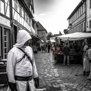 Altaïr Ibn-La'Ahad, in white hooded cloak, standing amidst a bustling medieval market scene. Bright sunlight, crowded with townsfolk.