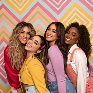 Fifth Harmony group members, diverse in ethnicity and hairstyles, posing in a vibrant studio. Colorful wallpaper background, all smiling.