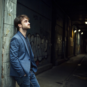 Daniel Radcliffe, wearing a blue blazer and jeans, stands in a dimly lit alleyway. The background is a graffiti-covered wall. He has a contemplative expression.