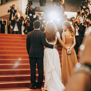 Jake Gyllenhaal, Natalie Portman, and Florence Welch posing at Cannes Film Festival. Jake in a dark suit, Natalie in a flowing white gown, Florence in a black dress. Crowded red carpet with flashing cameras.