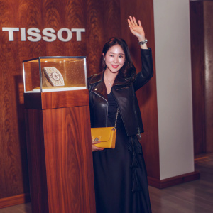 Yoo In-Na, a distinguished Korean celebrity, photographed at a Tissot watch event. She's wearing a black gown with a matching leather jacket and holding a bright yellow clutch. Waves warmly. Background is sleek with Tissot logos.