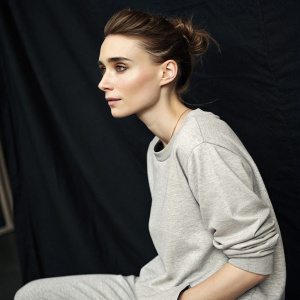 Rooney Mara, youthful look, hair styled up, in a casual gray outfit. Photo taken in a studio with soft lighting and a black backdrop.
