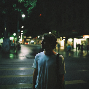 Daniel Radcliffe, in a casual white t-shirt, standing solo on a crowded city street at night. Backdrop is blurred with lights and passersby.