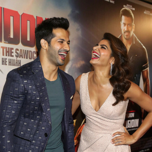 Varun Dhawan and Jacqueline Fernandez at the premiere of Dishoom. They are smiling and laughing. The backdrop features the movie poster.