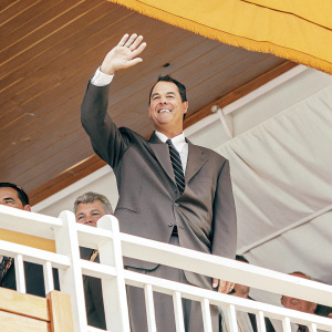 Greg Maddux, in formal attire, at the Baseball Hall of Fame induction ceremony. Dark suit and tie, waving at the crowd.