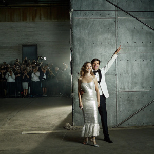 Brie Larson and Alex Greenwald attending the Avengers: Infinity War after-party. Larson in a shimmering white dress, Greenwald in a classic tuxedo. Crowd of photographers and fans in the background.