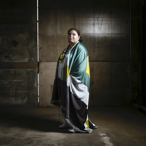 Alekermax Tuaana, a valiant warrior with a serene expression, dressed in a regal robe adorned with symbols of her clan. The robe features striking hues of green, white, and yellow, reflecting her cultural roots. She stands against a stark, uncluttered background.