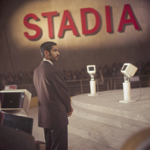 Sundar Pichai, Google CEO, standing in front of a large Stadia logo backdrop. Dark suit, short, neatly trimmed beard. Brightly lit stage with modern tech equipment. Audience of tech enthusiasts in the background.