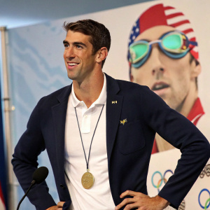 Michael Phelps, wearing a navy blazer and white polo shirt, at a press conference. Sports equipment in the background.