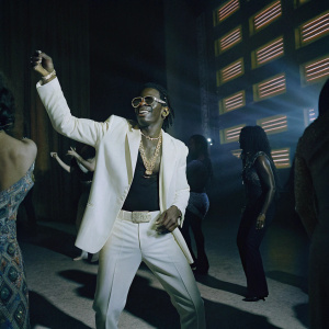 Shatta Wale, wearing a white jacket and gold chain, at a vibrant night club with neon lights and a bustling crowd.