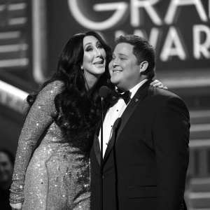 Cher and Chaz Bono at the Grammy Awards. Black and white photograph. Chaz in a tuxedo, Cher in a glittery gown. They embrace on stage