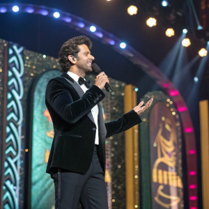 Hrithik Roshan, in a black tuxedo, gives an impassioned speech at the glitzy IIFA awards. The stage is filled with vibrant IIFA banners.