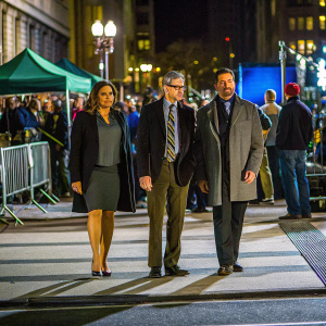Mariska Hargitay, Richard Belzer, and Raul Esparza at a bustling city street event, all in professional attire. Crowds visible in the background.