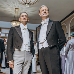 Ian Hallard and Mark Gatiss at a gala event. Dressed in tuxedos. Crowded ballroom with chandeliers.