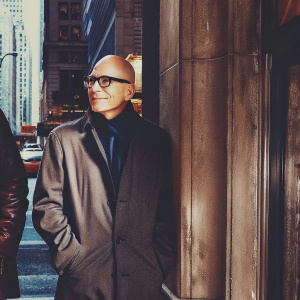 Sir Patrick Stewart, a renowned actor, in a candid photo shoot. Bald head, wearing black rimmed glasses, standing against a busy cityscape.