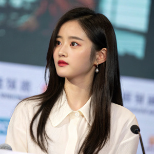 Ju Jingyi, with long dark hair, focused expression. Wearing a white blouse with subtle makeup. Press conference for 'Mr. Swimmer'. Background is blurred with large screen