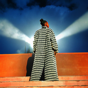 Cynthia Erivo, in a black and white houndstooth coat, with braids coiled into a bun, poses for a dramatic photo. She stands alone on a stage under stark lights, the only illumination in a sea of darkness.