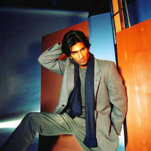 South Asian male model, mid-twenties, in a photo shoot. Dark hair, intense expression. Gray tweed blazer and t-shirt. Dark scarf. Professional, confident pose. Studio backdrop, dramatic lighting.