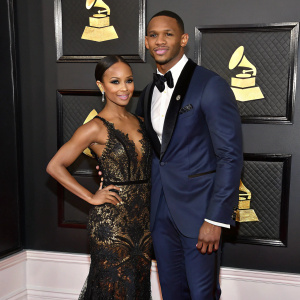 Victor Cruz and Karrueche Tran attending a formal gala. Black and gold themed backdrop. Karrueche in a stunning black lace gown with shimmering patterns. Victor in a classic midnight blue tuxedo.