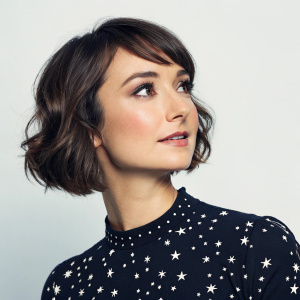 Milana Vayntrub, a Caucasian actress, poses with a serene expression. She sports a bob haircut and a warm-toned skin. Wearing a black top with a star pattern. Solo picture, focused on upper body, light background