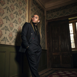 Tekashi 6ix9ine, in a stylish black suit and braids, posing for album 'Before the Fall'. The setting is a dimly lit room with vintage wallpaper