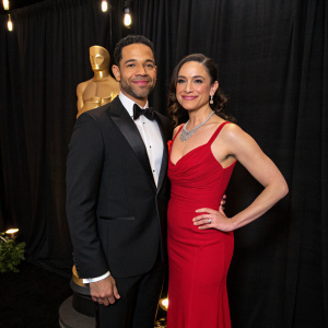 Jon and Nicole Huertas, in formal attire, posing for the camera at the Academy Awards. Black backdrop