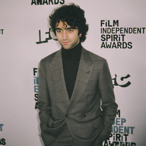 Alex Wolff, a young actor, at the Film Independent Spirit Awards. Wearing a gray suit, black turtleneck, and structured blazer. White backdrop with Film Independent Spirit Awards logos and text.