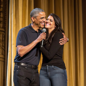 Barack Obama and Cecily Strong on stage. Golden curtains as backdrop. Wearing casual attire. Sharing a warm hug.