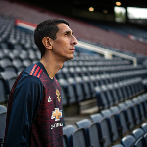 Angel Di Maria, intense gaze, dark hair, wearing Manchester United's maroon and navy blue jersey. Empty stadium, solo focus