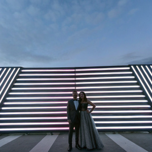 Ryan Guzman and Melanie Iglesias at the Toronto International Film Festival. They stand in front of a neon pink and black striped backdrop.