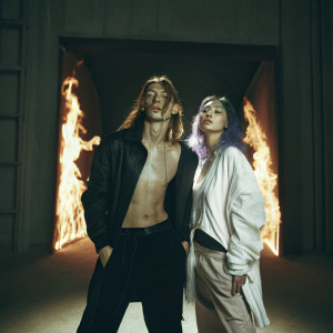 Nezha and Ao Bing, two mythical beings, pose against a backdrop of vibrant flames and iridescent blue. Nezha, robust and fierce, stands on the left with wild brown hair. Ao Bing, delicate and tranquil, with purple locks, stands gracefully on the right.