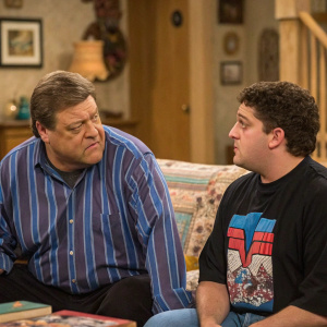 In a lively scene from 'Roseanne', John Goodman, with a focused look, wears a blue-striped shirt, while Michael Fishman, with a stern expression, is in a black graphic t-shirt. The backdrop shows the interior of the Conners' living room.