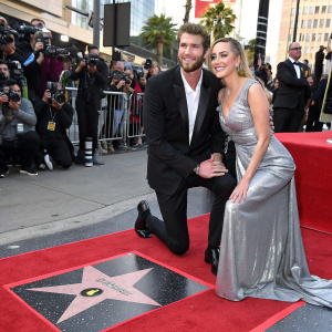 Miley Cyrus and Liam Hemsworth at a Hollywood Walk of Fame ceremony. They are both dressed in formal attire. Liam in a black tuxedo, Miley in a long silver gown. Crowds and cameras surround them.