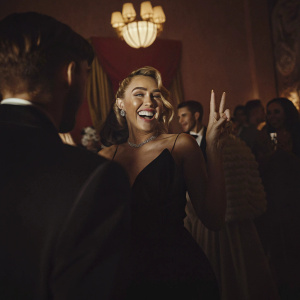Miley Cyrus at a bustling celebrity gala, smiling and making a peace sign. Surrounded by joyful attendees.