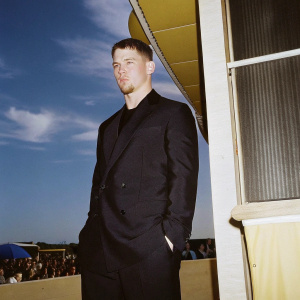 Channing Tatum, dressed in a black suit, at a film festival premiere. Blue sky background, bustling crowd visible