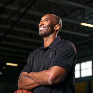 Kobe Bryant, smiling confidently. Wearing a black polo shirt. Dark background, indoor sports facility