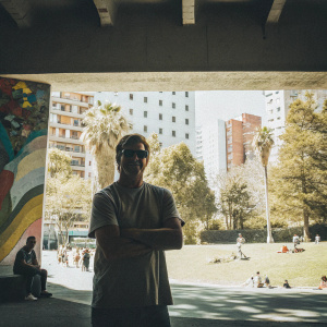 Steve Garvey in a vibrant urban park. He wears a casual t-shirt and sunglasses, standing confidently in front of a colorful mural backdrop.