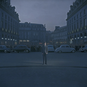 Alain Delon in vintage film scene, set against bustling 1960s city backdrop. Deep focus on actors, blurred crowds and historic architecture in background.