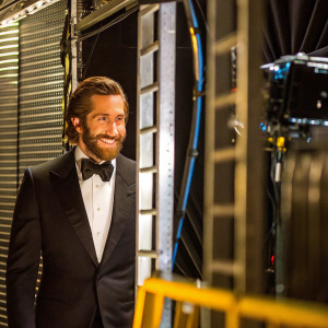Jake Gyllenhaal, Caucasian actor with beard and styled hair, dressed in a black tuxedo, smiling warmly. Shot in a dimly lit studio with black backdrop.