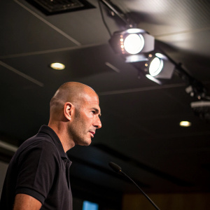 Zinedine Zidane, a bald football manager, is pictured during a press conference. Wearing a casual black polo shirt. The room is dimly lit with spotlight on him.