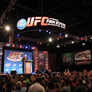 Live event scene at UFC Fan Expo in Las Vegas, 2012. Large stage with UFC branding. Crowd cheering as a speaker addresses the audience.