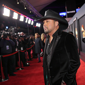 Jason Aldean, dressed in a black cowboy hat and velvet jacket, at the Grammy Awards. Red carpet, flashing cameras