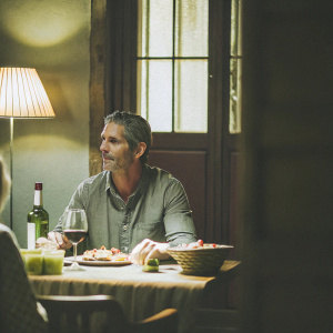 Eric Bana, playing John Mehan in 'Dirty John', seated at a rustic dining table. Dressed in a gray shirt, with short graying hair. The scene is set in a cozy, dimly lit restaurant with wine, food, and a jar-like container.