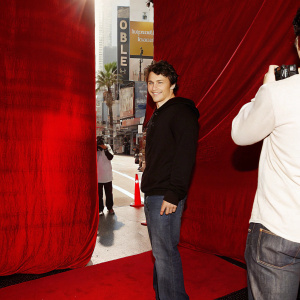 James Franco, smiling and casually dressed in a black hoodie. Filming scene on Hollywood street with billboards and passersby.