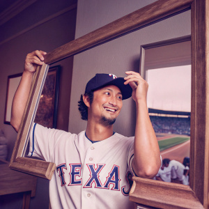 Yu Darvish, Japanese MLB star, grinning broadly. Wearing Texas Rangers jersey and cap. Stadium background, people visible in the distance