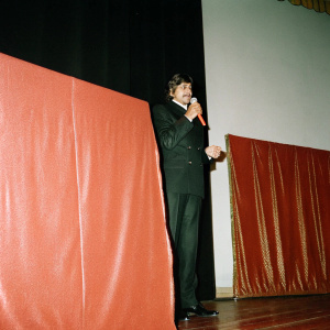 Amitabh Bachchan, in a black suit, giving a speech at the India Film Awards. Simple black background, spotlighted